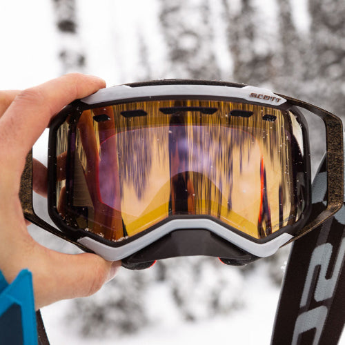 Close-up of a person holding a pair of goggles with a snowy background