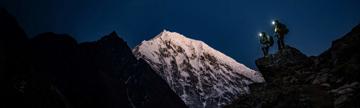 Two people with backpacks and headlamps standing on a rocky outcrop at night, silhouetted against a mountain peak.